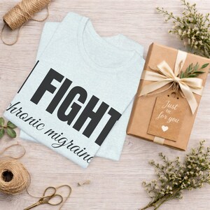 Puede incluir: Una camiseta azul claro con la palabra "FIGHT" en letras negras grandes y "chronic migraine" en texto cursivo m&aacute;s peque&ntilde;o. Una caja de regalo envuelta con una etiqueta que dice "Just for you" tambi&eacute;n est&aacute; en la imagen.