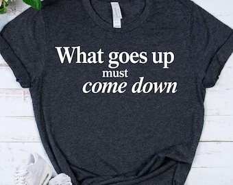 Puede incluir: Una camiseta de manga corta en color gris carbón oscuro con el texto impreso en blanco "What goes up must come down" centrado en el pecho. La prenda tiene cuello redondo y un corte estándar, con una etiqueta blanca visible en el cuello.