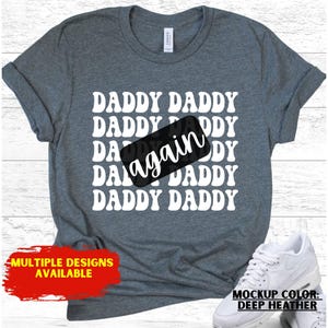 May include: Deep heather gray t-shirt with the words "DADDY" repeated multiple times in white, with the word "again" in a black rectangle. The shirt has rolled-up sleeves and a white tag. The text "MULTIPLE DESIGNS AVAILABLE" is in a red rectangle.