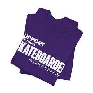 以下が含まれることがあります： SUPPORT YOUR LOCAL SKATEBOARDER BY DRAINING YOUR POOLという白いテキストがプリントされた紫色のTシャツ。クルーネックと半袖のデザインで、胸元には太字のサンセリフ体でグラフィックが配置されています。