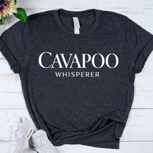 May include: A dark heather gray short-sleeve t-shirt featuring the white text CAVAPOO WHISPERER printed in a bold, serif font across the chest. The garment has a crew neckline and a standard fit, with a white label visible at the inner collar.