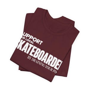 以下が含まれることがあります： SUPPORT YOUR LOCAL SKATEBOARDER BY DRAINING YOUR POOLという白い文字がプリントされた、折りたたまれたマルーンカラーのTシャツ。クルーネックと半袖のデザインで、胸元には太字のフォントでグラフィックが配置されています。