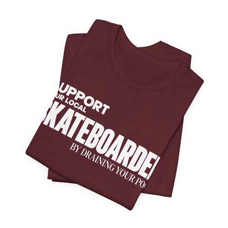 以下が含まれることがあります： SUPPORT YOUR LOCAL SKATEBOARDER BY DRAINING YOUR POOLという白い文字がプリントされた、折りたたまれたマルーンカラーのTシャツ。クルーネックと半袖のデザインで、胸元には太字のフォントでグラフィックが配置されています。