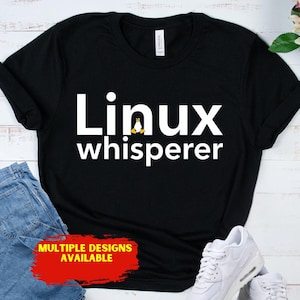 May include: Black t-shirt with the text "Linux whisperer" in white, featuring a penguin graphic. The shirt is displayed with blue jeans and white sneakers. A red banner at the bottom reads "MULTIPLE DESIGNS AVAILABLE".