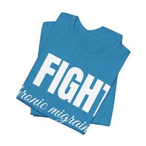 Puede incluir: Una camiseta turquesa doblada con la palabra "FIGHT" en letras blancas grandes. Debajo, en una fuente cursiva m&aacute;s peque&ntilde;a, el texto dice "chronic migraine". La camiseta est&aacute; hecha de un material suave.