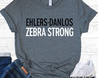 May include: A deep heather grey t-shirt featuring the text "EHLERS-DANLOS ZEBRA STRONG" in black and white. The shirt has a crew neck and rolled sleeves. The mockup colour is deep heather.