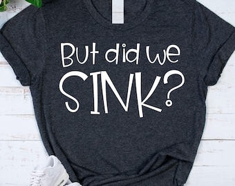 May include: Dark gray t-shirt with the white text "But did we SINK?". The shirt has a v-neck and rolled-up sleeves. White sneakers are visible in the bottom left corner. The shirt is likely made of a soft, comfortable material.