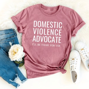 Op de afbeelding: Een roze T-shirt met de tekst "Domestic Violence Advocate I'll be there for you" in witte letters.