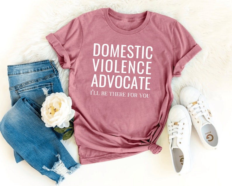 Op de afbeelding: Een roze T-shirt met de tekst "Domestic Violence Advocate I'll be there for you" in witte letters.