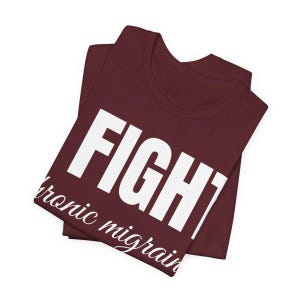 Puede incluir: Camiseta burdeos doblada con la palabra "FIGHT" en letras blancas grandes y las palabras "chronic migraine" en letras blancas m&aacute;s peque&ntilde;as. La camiseta es de un material suave.