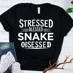 May include: Black t-shirt with the white text "STRESSED BLESSED SNAKE OBSESSED". The shirt is displayed with blue jeans and white sneakers. The shirt is made of a soft material.