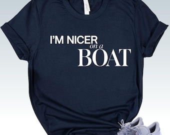 May include: A dark navy blue short-sleeved t-shirt featuring the white text "I'M NICER on a BOAT" printed across the chest. The shirt has a crew neckline and rolled sleeves, paired with a pair of light gray athletic sneakers with white laces.