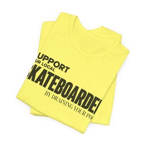 以下が含まれることがあります： SUPPORT YOUR LOCAL SKATEBOARDER BY DRAINING YOUR POOLという太字の黒いテキストがプリントされた、明るい黄色のTシャツ。クルーネックと半袖のデザインで、グラフィックがはっきりと見えるように折りたたまれた状態で配置されています。