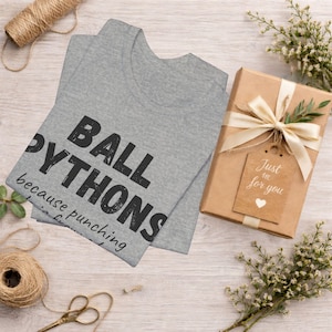 May include: A gray t-shirt with the text "BALL PYTHONS" in large black letters. The shirt is folded and next to a wrapped gift box with a tag that reads "Just for you". Decorative elements include twine, scissors, and flowers.