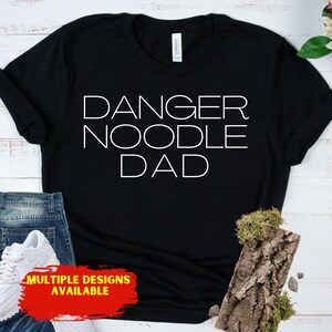 May include: Black t-shirt with the white text "DANGER NOODLE DAD". The shirt has a crew neck and short sleeves. The bottom of the shirt is partially visible, along with a pair of blue jeans and a white sneaker. A red and yellow rectangle at the bottom says "MULTIPLE DESIGNS AVAILABLE".