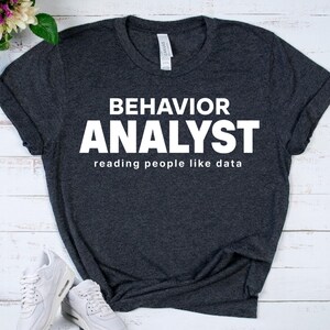 May include: Dark gray t-shirt with the words "BEHAVIOR ANALYST" in large white letters. Below, the text reads "reading people like data." The shirt is folded, with white sneakers visible in the lower left corner. The shirt is made of a soft material.
