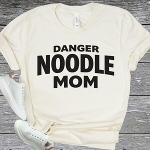 May include: Cream-colored t-shirt with the text "DANGER NOODLE MOM" in bold black letters. The shirt has a classic crew neck and short sleeves. White sneakers are visible in the lower left corner.