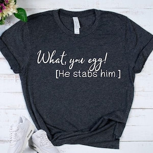 May include: Dark gray t-shirt with the text "What, you egg! [He stabs him.]" in white script. The shirt is a crew neck style and has short sleeves. White sneakers are visible in the bottom left corner.