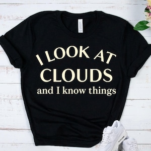 May include: Black t-shirt with the text "I LOOK AT CLOUDS and I know things" in a cream-colored font. The shirt has rolled-up sleeves and a tag that says "CANVAS". White sneakers are visible in the bottom right corner.
