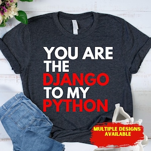 May include: Dark gray t-shirt with the text "YOU ARE THE DJANGO TO MY PYTHON" in white and red. The shirt is folded next to a pair of blue jeans and white sneakers. The phrase "MULTIPLE DESIGNS AVAILABLE" is in a red box.