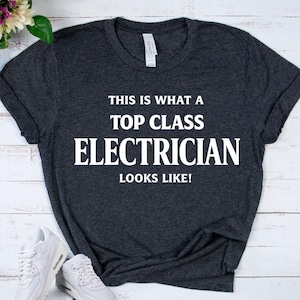 May include: A dark heather gray short-sleeved t-shirt with white text printed on the front that reads: THIS IS WHAT A TOP CLASS ELECTRICIAN LOOKS LIKE! The shirt features a crew neckline and a standard fit, showcasing a clean and simple design.