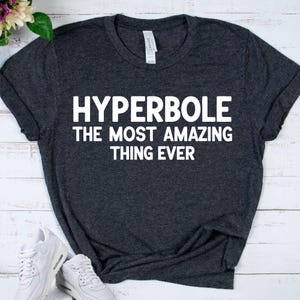 May include: Dark gray t-shirt with the white text "HYPERBOLE THE MOST AMAZING THING EVER." The shirt is folded, with white sneakers visible in the lower left corner. The shirt has a tag that says "CANVAS".