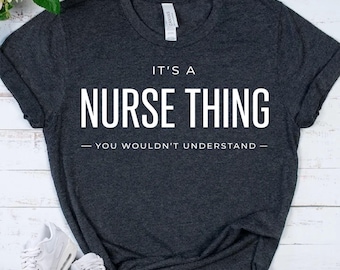 Puede incluir: Una camiseta de manga corta en color gris marengo oscuro con el texto blanco "IT'S A NURSE THING - YOU WOULDN'T UNDERSTAND" impreso en el pecho. La prenda tiene cuello redondo y un corte estándar, presentada sobre una superficie plana.