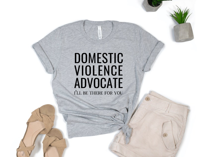 Op de afbeelding: Grijs T-shirt met de tekst "Domestic Violence Advocate" en "I'll be there for you" in zwart.