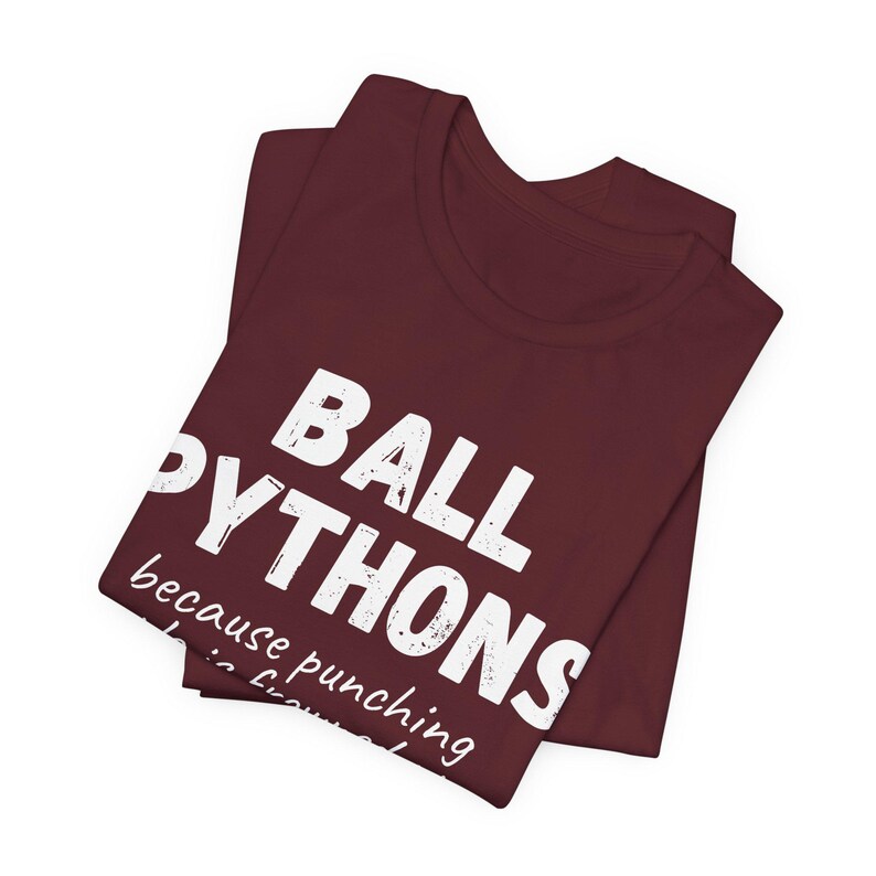 May include: A folded maroon t-shirt with the words "BALL PYTHONS" in large white letters. Below, smaller text reads "because punching is frowned upon." The shirt is made of soft material.