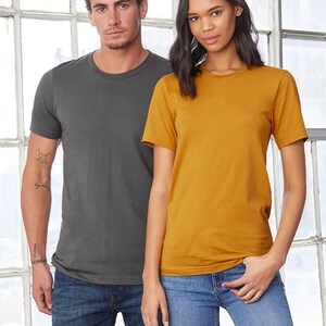Op de afbeelding: Twee mensen dragen T-shirts, een in antracietgrijs en de andere in mosterdgeel. Beide dragen een blauwe jeans.