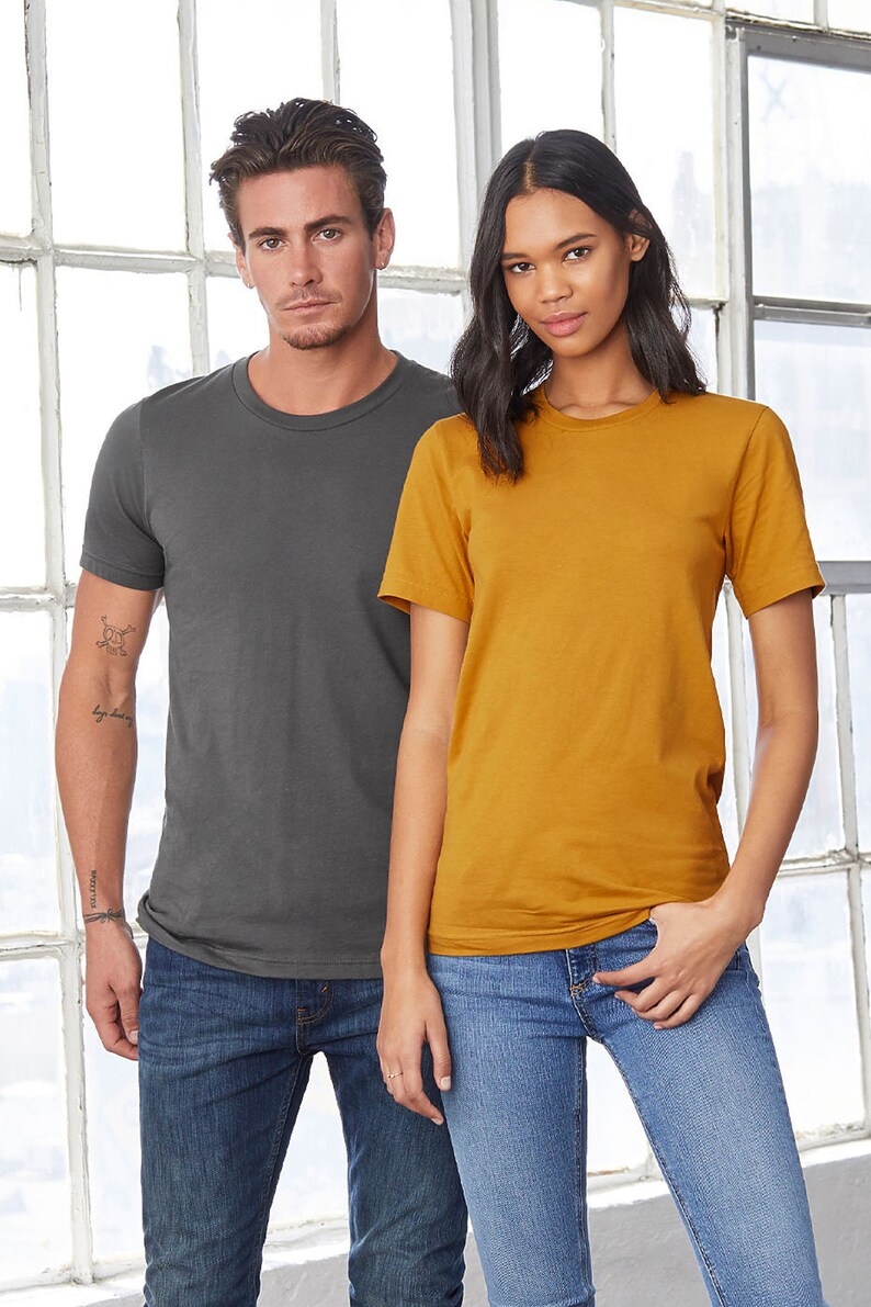 Op de afbeelding: Twee mensen dragen T-shirts, een in antracietgrijs en de andere in mosterdgeel. Beide dragen een blauwe jeans.