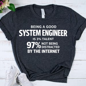 May include: Dark gray t-shirt with white text that reads "BEING A GOOD SYSTEM ENGINEER IS 3% TALENT 97% NOT BEING DISTRACTED BY THE INTERNET." The shirt has a rolled sleeve and a tag that says "CANVAS."