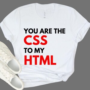 May include: White t-shirt with the text "YOU ARE THE CSS TO MY HTML" in black and red. The shirt is paired with white sneakers. The shirt is made of a soft material.