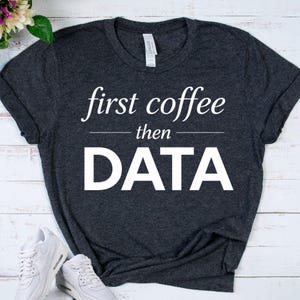 May include: Dark gray t-shirt with the text "first coffee then DATA" in white. The shirt is a crew neck style and appears to be made of a soft material. White sneakers are visible in the bottom of the image.