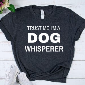 May include: A dark heather gray short-sleeve t-shirt featuring the white printed text TRUST ME I'M A DOG WHISPERER. The shirt has a crew neckline and a standard fit, with the text centered prominently on the front of the garment.