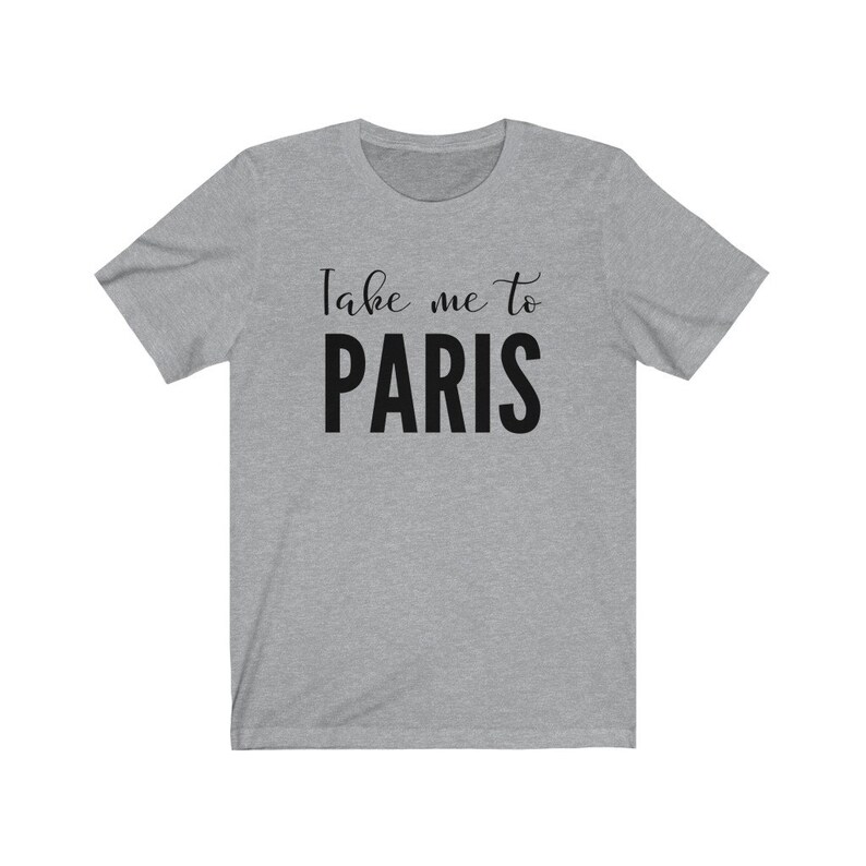 May include: A heather gray short-sleeve t-shirt featuring the text Take me to PARIS printed in black across the chest. The words Take me to are in a cursive script font, while the word PARIS is in a bold, uppercase sans-serif typeface.