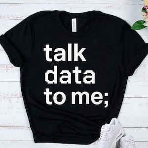 May include: A black short-sleeved t-shirt featuring the white pixelated text "talk data to me;" printed on the front. The shirt has a crew neckline and a standard fit, with a white brand label visible inside the collar area at the back neckline.