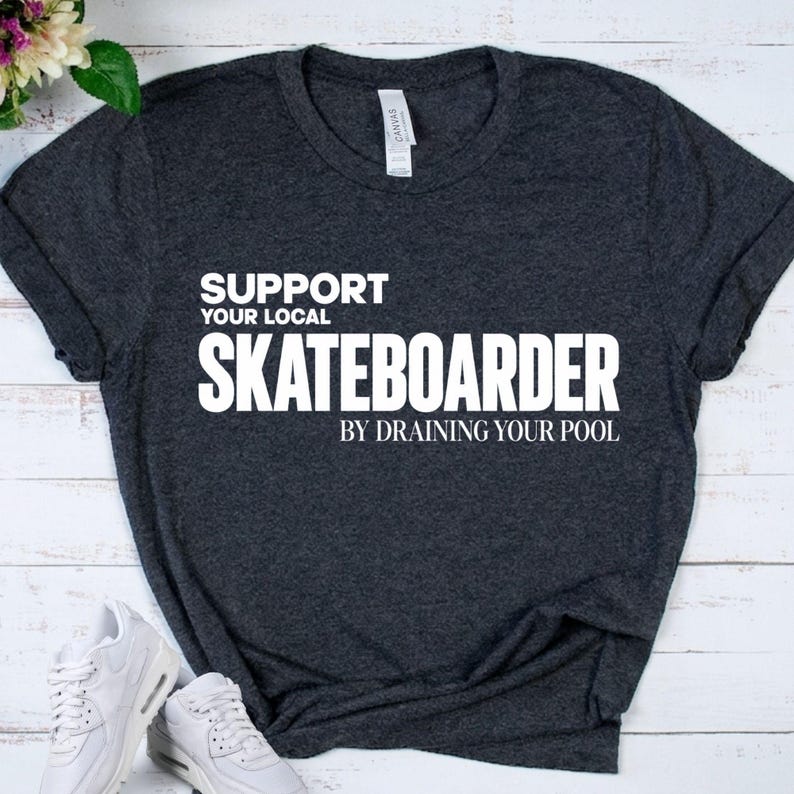 以下が含まれることがあります： ダークヘザーグレーの半袖Tシャツで、前面に「SUPPORT YOUR LOCAL SKATEBOARDER BY DRAINING YOUR POOL」という白い文字がプリントされています。クルーネックの標準的なフィット感で、襟の内側には白いブランドタグが見えています。