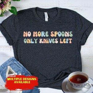 May include: Dark gray t-shirt with the text "NO MORE SPOONS ONLY KNIVES LEFT" in pastel colors. The shirt is paired with blue jeans, a cup of tea, and a floral arrangement. The words "MULTIPLE DESIGNS AVAILABLE" are also visible.