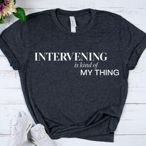 May include: A dark charcoal gray short-sleeve t-shirt featuring the white printed text INTERVENING is kind of MY THING centered on the chest. The shirt has a crew neckline and a standard fit, with a visible brand label inside the collar area.