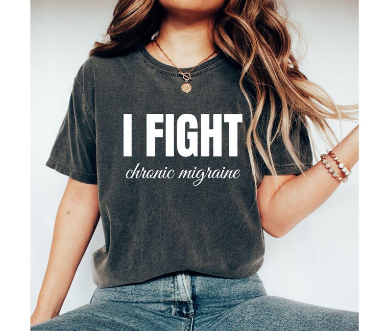 May include: A dark gray, short-sleeved t-shirt features the text 'I FIGHT chronic migraine' in white lettering. The shirt is styled on a model. The text is bold and simple, with the words 'I FIGHT' stacked above the phrase 'chronic migraine' in a more stylized script font. The shirt appears to be made of a soft, comfortable fabric with a slightly distressed or vintage look.
