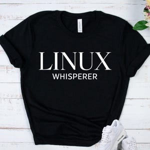 May include: A black short-sleeved t-shirt featuring the white text LINUX WHISPERER printed in a bold serif font across the chest. The garment has a crew neckline and a standard fit, with a white brand label visible inside the collar area.