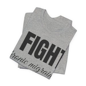 Puede incluir: Una camiseta gris doblada con la palabra "FIGHT" en letras negras grandes y en negrita. Debajo de la palabra "FIGHT" est&aacute; el texto "chronic migraines" en una fuente cursiva m&aacute;s peque&ntilde;a. La camiseta est&aacute; hecha de un material suave.