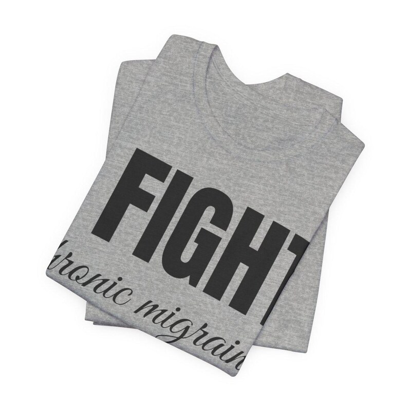 Puede incluir: Una camiseta gris doblada con la palabra "FIGHT" en letras negras grandes y en negrita. Debajo de la palabra "FIGHT" est&aacute; el texto "chronic migraines" en una fuente cursiva m&aacute;s peque&ntilde;a. La camiseta est&aacute; hecha de un material suave.