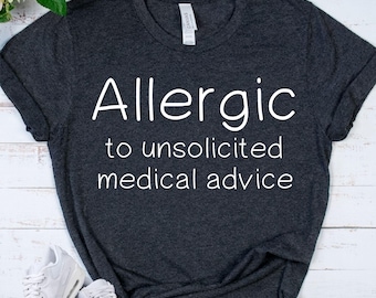 May include: Dark grey t-shirt with the white text "Allergic to unsolicited medical advice." The shirt is a crew neck style and appears to be made of a soft material. The shirt is displayed on a light-coloured surface.