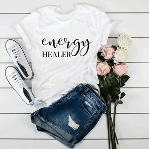 Pode incluir: Camiseta branca com o texto "energy healer" em letras de escrita preta. A camiseta est&aacute; colocada sobre uma superf&iacute;cie de madeira branca com um par de t&ecirc;nis brancos e um buqu&ecirc; de rosas cor-de-rosa.