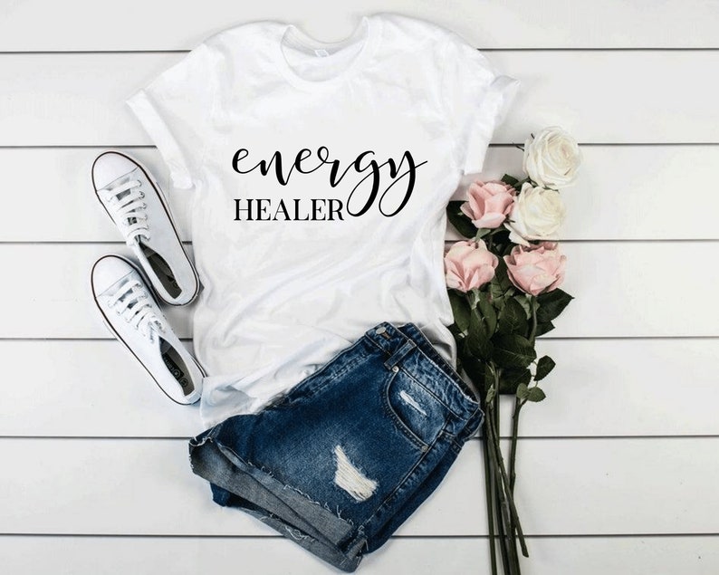 Pode incluir: Camiseta branca com o texto "energy healer" em letras de escrita preta. A camiseta est&aacute; colocada sobre uma superf&iacute;cie de madeira branca com um par de t&ecirc;nis brancos e um buqu&ecirc; de rosas cor-de-rosa.