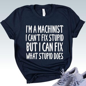 May include: Navy blue t-shirt with the text "I'M A MACHINIST I CAN'T FIX STUPID BUT I CAN FIX WHAT STUPID DOES" in white. The shirt is folded with a pair of gray sneakers in the bottom right corner.