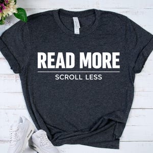 May include: Dark gray t-shirt with the white text "READ MORE" above "SCROLL LESS". The shirt is made of a soft material and has short sleeves. White sneakers are visible in the bottom left corner.