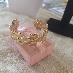 Puede incluir: Una pulsera dorada con un diseño floral, apoyada sobre una pequeña caja de regalo rosa. La pulsera presenta una serie de elementos en forma de flor. La caja está atada con una cinta blanca. El fondo es una superficie suave y texturizada.